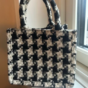 Svartvit rutig handväska från H&M - Snygg handväska från H&M i svartvitt rutigt mönster. Väskan har två handtag och en dragkedja för säker förvaring. Perfekt för att ge din outfit en stilren touch.