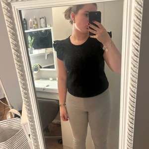 Svart topp med volangdetaljer - Säljer en stilren svart topp med volangdetaljer vid ärmarna. Toppen har en rund halsringning och är kortärmad, perfekt för både vardag och festliga tillfällen.