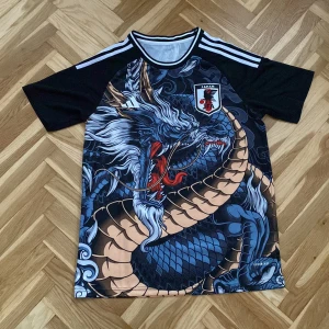 Svart och blå fotbollströja med drakmotiv  - Cool fotbollströja från Adidas och Japan med ett imponerande drakmotiv i blått och beige. Tröjan har korta ärmar och klassiska Adidas-ränder på axlarna. Perfekt för dig som vill sticka ut på planen!