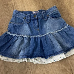 Blå jeanskjol med volang - Jeans kjol från Joelle i storlek 36 med volang och blommigt tyg. Kjolen är 35cm lång och är 37cm i midjan(rakt över).