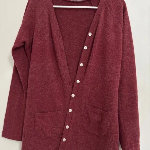Vinröd kofta med knappar - Mysig vinröd kofta med knappar framtill och två praktiska fickor. Perfekt för kyliga dagar och att ha som ett extra lager. Långärmad design som ger en avslappnad och bekväm stil.