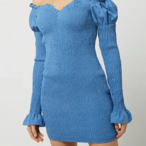 Blå off-shoulder klänning från Sthlm Blvd - Snygg blå off-shoulder klänning från Sthlm Blvd med smockad design och långa ärmar. Perfekt för en stilren look med en touch av elegans. Klänningen har en åtsittande passform som framhäver figuren.