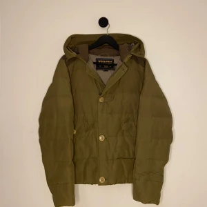 Woolrich jacka  - Säljer en olivgrön dunjacka från Woolrich med huva och knappar framtill. Jackan har en quiltad design och praktiska fickor med lock. Perfekt för kyliga dagar. Hör gärna av er vid flera frågor eller funderingar! 