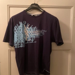 Mörkblå t-shirt från Yves Saint Laurent - Snygg mörkblå t-shirt från Yves Saint Laurent med ett coolt tryck i ljusblått och vitt på framsidan. Perfekt för en stilren look. T-shirten har korta ärmar och rund halsringning. Det står XL i den men tröjan är vintage så det passar M-L. 