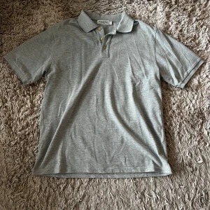 Yves Saint Laurent Piké - Säljer en grå pikétröja från Yves Saint Laurent. Tröjan har korta ärmar och en klassisk krage med tre knappar. Den är broderad med YSL-loggan på bröstet. Perfekt för en avslappnad men ändå elegant look. Lappen vid nacken är av på ena sidan, går däremot att laga. Därav är priset lägre. Skriv vid frågor!