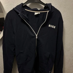 Svart hoodie från BOSS - Säljer en stilren svart hoodie från BOSS med dragkedja och huva. Tröjan har långa ärmar och en vit logotyp på bröstet. Perfekt för en avslappnad look.