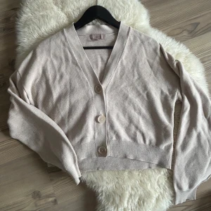 Beige kofta med knappar - Säljer en beige kofta med stora knappar framtill. Den är croppad och har en v-ringad design med långa ärmar. Perfekt för en stilren och avslappnad look.