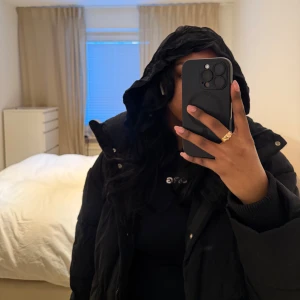 Svart pufferjacka från Zara - Säljer en svart pufferjacka från Zara med huva och dragkedja. Jackan har en quiltad design och knappar framtill. Perfekt för kyliga dagar.