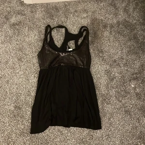 Svart paljettlinne - Snyggt svart linne med paljettdetaljer upptill. Perfekt för en kväll ute eller en festlig tillställning. Linnet har en racerback-design och är tillverkat i ett mjukt material som ger en bekväm passform.