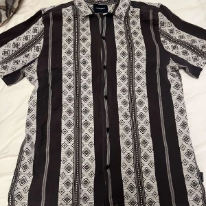 Svart och vit mönstrad skjorta från Lindbergh  - Snygg kortärmad skjorta från Lindbergh med ett unikt svart och vitt mönster. Skjortan har knappar framtill och en klassisk krage. Perfekt för en avslappnad stil.