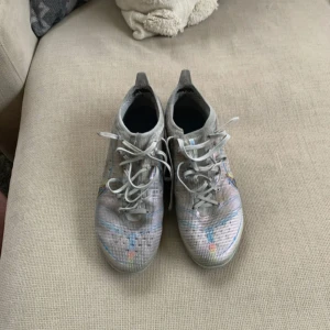 Nike fotbollsskor med färgglada detaljer - Snygga fotbollsskor från Nike i en ljusgrå nyans med färgglada detaljer och snörning. Skorna har en modern design med en tydlig Nike-logga på sidan. Perfekta för fotbollsplanen!