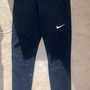 Svarta träningsbyxor från Nike - Säljer ett par svarta träningsbyxor från Nike med en vit logga på benet. Byxorna har en elastisk midja och är perfekta för träning. De är tillverkade i ett mjukt och stretchigt material för bästa komfort.