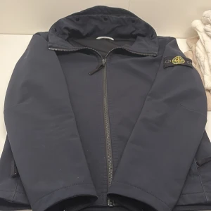 Svart jacka från Stone Island - Snygg mörkblå/svarr jacka från Stone Island med dragkedja. Jackan har en ikonisk logotyp på ärmen och är perfekt för kyligare dagar. Den har en stilren design som passar till många olika outfits. Hör av dig vid intresse!!