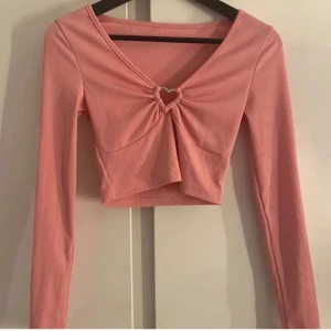 Rosa ribbad croppad topp från Shein - Säljer en söt rosa ribbad topp från Shein. Den är croppad med en v-ringning och har en knutdetalj framtill. Toppen har långa ärmar och är perfekt för en trendig look.