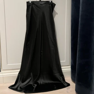 Lång svart kjol i satin NA-KD - Superfin svart långkjol i satin från NA-KD! I storlek 32 med en diskret dragkedja på sidan. Den mäter 95cm lång. Perfekt till både vardags och fest, enkel att matcha med olika toppar och accessoarer🖤😇 Kjolen är i nyskick med lappen kvar! 