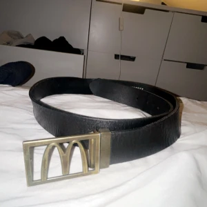 Mcdonalds bälte - Snyggt svart bälte med ett unikt guldfärgat spänne som har en distinkt design. Perfekt för att ge en stilren touch till din outfit. Mcdonalds.