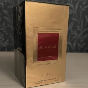 Bella Rouge Eau de Parfum från Riiffs - Upptäck Bella Rouge Eau de Parfum från Riiffs, en lyxig doft i en elegant guld- och röd förpackning. Flaskan rymmer 100 ml och är perfekt för den som söker en sofistikerad och förförisk doftupplevelse. Parfymen är fortfarande inplastad och oanvänd. 