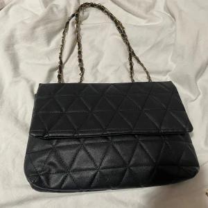 Svart quiltad axelväska - Snygg svart quiltad axelväska med kedjehandtag. Perfekt för att ge en elegant touch till din outfit. Väskan har en stilren design och är tillverkad i syntetmaterial.