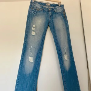 Blå lågmidjade bootcut jeans med nitar - Snygga blå jeans med slitna detaljer och coola nitar vid fickorna. De är bootcut och lågmidjade. Jeansen har en klassisk femficksdesign och knappgylf. Sjukt snygga❤️