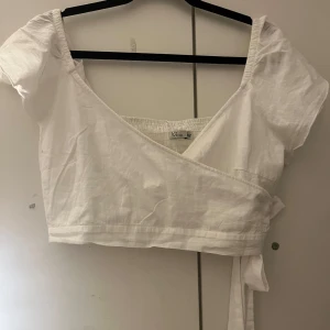 Vit omlottopp från NLY Trend - Snygg vit omlottopp från NLY Trend med korta ärmar och knytband i midjan. Perfekt för en somrig look. Toppen har en smockad rygg för extra komfort och stil.