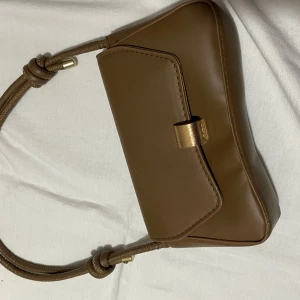 Brun axelväska med gulddetalj - Snygg brun axelväska i skinn med en elegant gulddetalj på framsidan. Väskan har en stilren design och en praktisk axelrem som gör den lätt att bära. Perfekt för att ge din outfit en touch av klass.