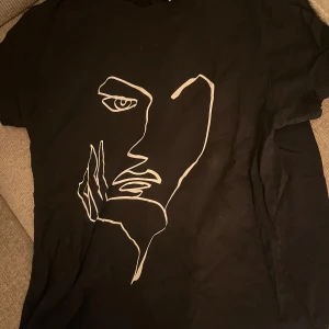 Svart t-shirt med ansiktsmotiv från Visual Clothing Project - Snygg svart t-shirt från Visual Clothing Project med ett stilrent ansiktsmotiv i vitt på framsidan. Perfekt för en casual look.