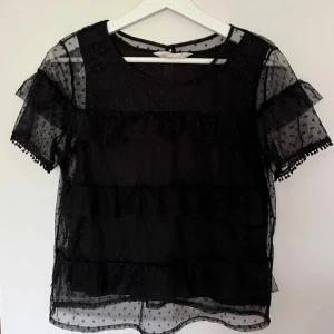Svart  blus med volanger - Snygg svart blus i transparent material med prickigt mönster. Blusen har korta ärmar och dekorativa volanger som ger en trendig look. Perfekt för en kväll ute eller en speciell tillställning.