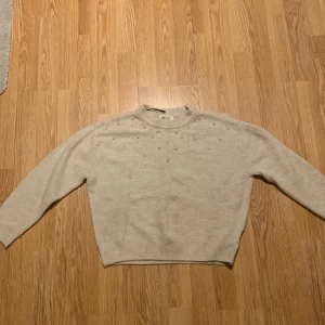 Beige tröja med pärlor från H&M - Säljer en söt beige tröja från H&M dekorerad med pärlor över bröstet. Tröjan har långa ärmar och en rund halsringning. Perfekt för en stilren och mysig look.
