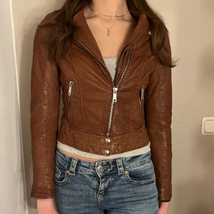 Brun skinnjacka från Jofama - Snygg brun skinnjacka från Jofama designad av Marie Serneholt. Jackan har en dragkedja framtill och stiliga detaljer med knappar. Perfekt för en cool och trendig look.