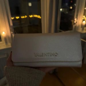 Campanjefärgad väska från Valentino - Elegant champagnje färgad väska från Valentino med flera fack för kort och en praktisk dragkedja inuti. Perfekt för att hålla ordning på dina tillhörigheter med stil. Kommer i originalförpackning. Den är äkta, endast använd en eftermiddag