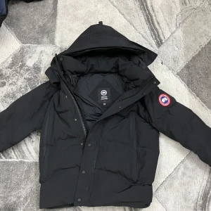 Svart dunjacka från Canada Goose - Säljer en svart dunjacka från Canada Goose med avtagbar huva och pälsdetalj. Jackan har en klassisk design med dragkedja och knappar framtill samt en ikonisk logga på ärmen. Perfekt för kalla vinterdagar. (Tar emot byten bara)