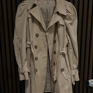 Beige trenchcoat från Gina Tricot - Snygg beige trenchcoat från Gina Tricot med dubbelknäppning och puffiga axlar. Jackan har långa ärmar och ett bälte i midjan för en stilren look. Perfekt för vår och höst!