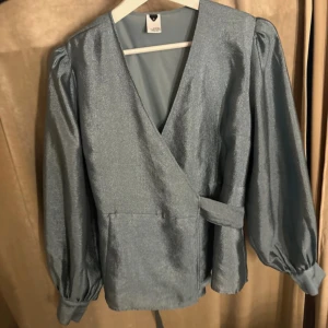 Glittrig omlottopp i blått - Snygg glittrig omlottopp från MQ i en vacker blå nyans. Toppen har långa ärmar med puffiga axlar och ett knytband i midjan för en smickrande passform. Perfekt för en festlig kväll! Storlek S