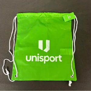 Grön gympapåse från Unisport - Säljer en grön gympapåse från Unisport med vitt tryck och snören. Perfekt för att bära träningskläder eller andra småsaker. Enkel och praktisk design.