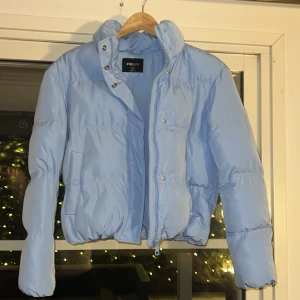 Ljusblå pufferjacka från New Yorker - Säljer en snygg ljusblå pufferjacka från NewYorker. Jackan har en hög krage och stängs med både dragkedja och knappar. Perfekt för kyliga dagar med sin vadderade design. Det är i storlek S