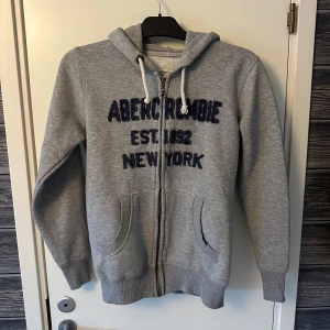 Grå hoodie från Abercrombie & Fitch - Säljer en grå hoodie från Abercrombie & Fitch med dragkedja och broderad text i marinblått på framsidan. Tröjan har en mysig huva med dragsko och två fickor framtill. 