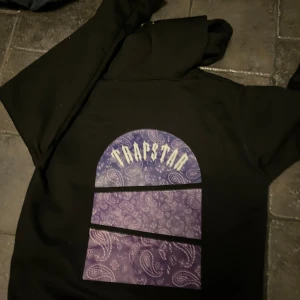 Svart hoodie från Trapstar - Snygg svart hoodie från Trapstar med lila paisley-mönster och logga på både fram- och baksidan. Perfekt för en avslappnad stil med en touch av streetwear.