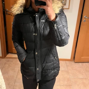 Svart dunjacka från Rockandblue -  svart dunjacka från Rockandblue med pälskantad huva. Jackan har dragkedja och knappar framtill samt praktiska fickor. Perfekt för kyliga dagar. Stories 38. Knappt använd 