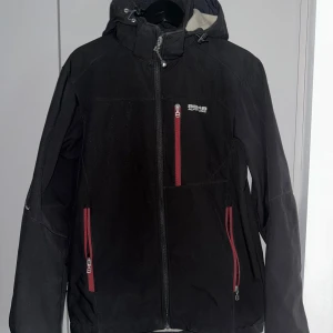 Svart jacka från 8848 - Snygg svart jacka från 8848 med röda dragkedjor och justerbar huva. Perfekt som skaljacka kyligare dagar med sin funktionella design och praktiska fickor. 