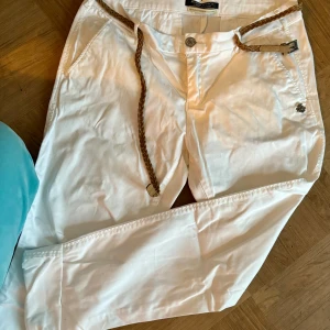 Vita byxor från Maison Scotch - Snygga vita byxor från Maison Scotch med flätat bälte. Byxorna har en normal passform och är perfekta för en stilren look. De har knappar och dragkedja framtill samt fickor både fram och bak.