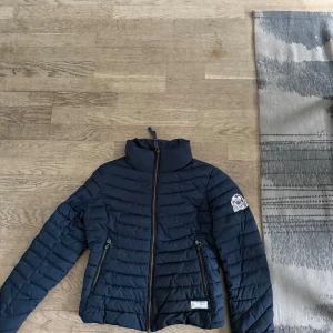 Mörkblå pufferjacka med dragkedja - Snygg mörkblå pufferjacka med dragkedja och hög krage. Jackan har ett märke på ärmen och praktiska fickor med dragkedjor. Perfekt för kyligare dagar.