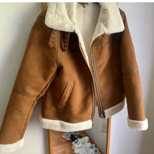 Brun mockajacka med foder - Snygg brun mockajacka med vit fuskpälsfoder och dragkedja. Jackan har en liten defekt på ena ärmen (dm för bild) 