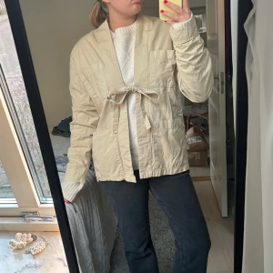 Beige jacka med knytband - Säljer en stilren beige jacka med knytband framtill. Jackan har en avslappnad passform och långa ärmar, perfekt för vår och höst. Enkel och chic design som passar till många olika outfits.