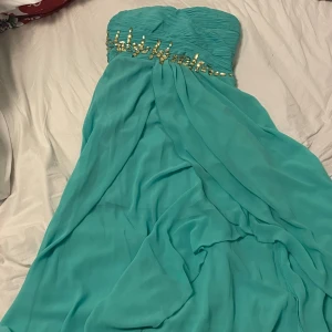 Turkos axelbandslös klänning  - Säljer en elegant turkos axelbandslös klänning från. Klänningen har vackra guldfärgade detaljer vid bysten och ett draperat fall. Perfekt för speciella tillfällen. Köpt i andra hand, fel storlek. Bra skick!