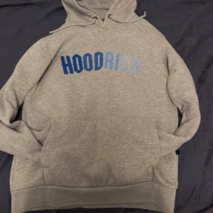 Grå hoodie från Hoodrich - Säljer en grå hoodie från Hoodrich med blå text på bröstet. Den har en klassisk design med känguruficka och justerbar huva. Perfekt för en avslappnad stil.