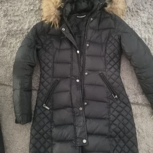 Svart dunjacka från Rockandblue - Snygg svart dunjacka från Rockandblue med quiltade detaljer och en mysig pälsfodrad huva. Jackan har dragkedja och knappar framtill samt praktiska fickor med dragkedja. Perfekt för kalla dagar! Har ett litet hål men de går att sy änkelt köpt för 3500 kr 