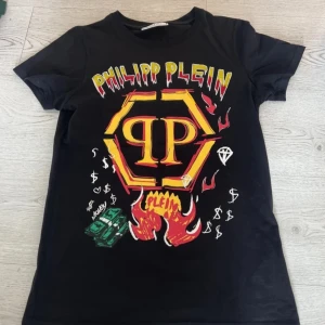 Svart t-shirt från Philipp Plein Junior - Cool svart t-shirt från Philipp Plein Junior med färgglatt tryck och logotyp på framsidan. Designen inkluderar eldflammor, diamanter och dollartecken. Perfekt för en ungdom som vill sticka ut med stil.