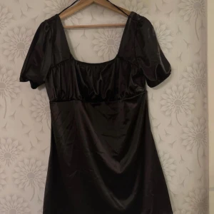 Svart klänning med puffärmar - Elegant svart klänning med korta puffärmar och fyrkantig urringning. Tillverkad i ett glansigt material som ger en lyxig känsla. Perfekt för en stilren look.