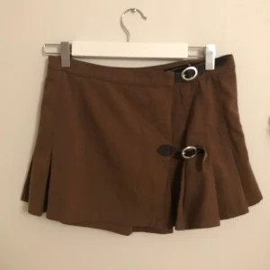 Brandy Melville brun kortkjol med spänne - Snygg brun kortkjol från brandy melville med omlott-design och dekorativa spännen framtill. Kjolen har en lätt utställd form som ger en fin siluett.