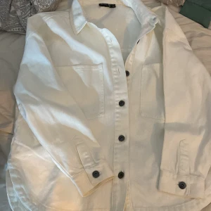Vit overshirt med knappar - Säljer en stilren vit overshirt med svarta knappar. Den har två praktiska bröstfickor och är perfekt för lager-på-lager-stil. Passar både till vardags och lite mer uppklädda tillfällen. Skjortan är i bra skick och redo för nya äventyr!Strl 42
